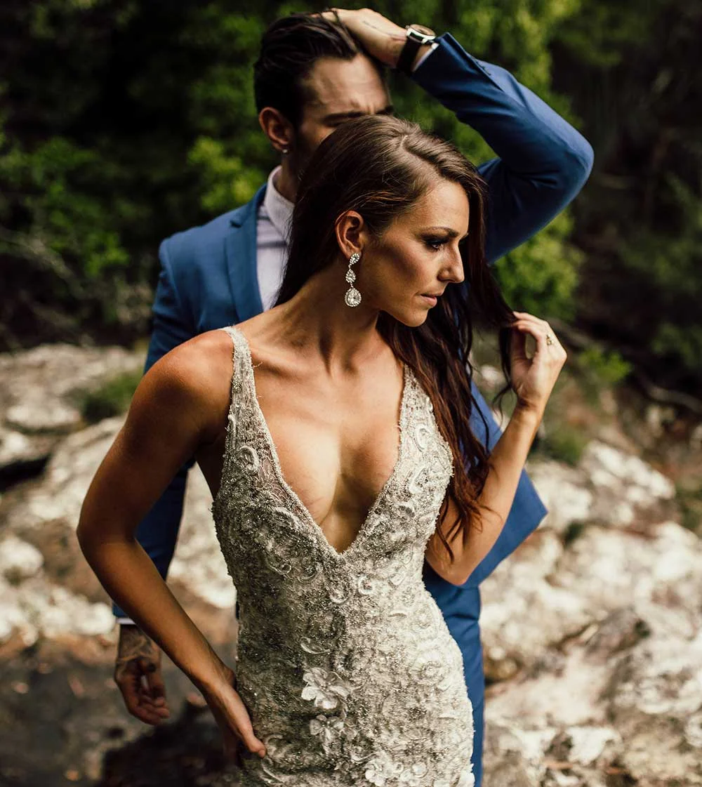 A woman in a sequined, form-fitting gown with a deep V-neckline standing in front of a man wearing a blue suit, with his hand resting on his own head.