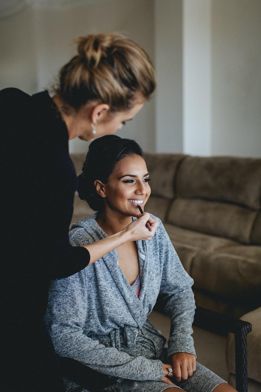 A makeup artist is shown applying lip liner to a smiling woman who is dressed in a cozy, grey robe.